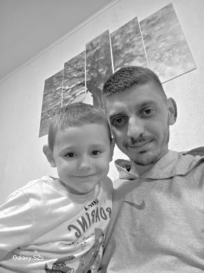 A black and white image displays two individuals, an adult male and a young male child, positioned in a close-up, selfie-style shot indoors in Karnalovo, Bulgaria. Both subjects face the camera directly with smiles. The adult male, situated on the right, exhibits short, dark hair and facial stubble, and wears a light-toned hooded sweatshirt featuring a distinct Nike swoosh logo on the chest. The child, positioned on the left, has short, light-toned hair and is dressed in a light-colored long-sleeved shirt with inverted text and graphical elements across the front. The background consists of a plain light-colored wall adorned with a four-panel grayscale artwork depicting a tree or a natural landscape. A segment of a ceiling and its decorative molding are visible in the upper left quadrant of the frame. The bottom left corner of the image contains a "Galaxy S25" watermark.