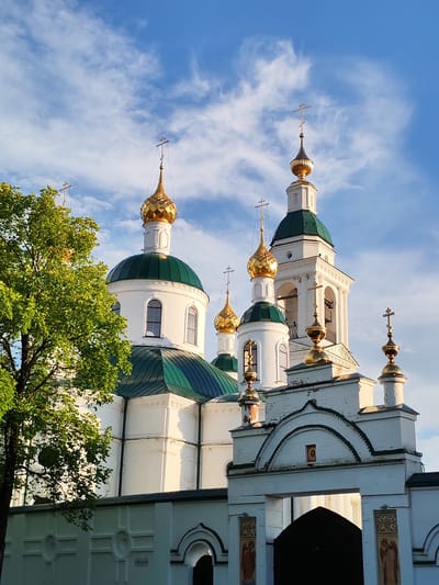 This is a photograph of the Church of the Resurrection in Chelganovo, Russia. The image captures the church on a clear day with a bright blue sky dotted with scattered white clouds, suggesting it was taken during daylight hours, likely in the late afternoon given the warm lighting.
The main subject is the church itself, a predominantly white building with striking green domes and ornate golden cupolas topped with crosses. The architecture is traditional Orthodox, with multiple domes of varying sizes and a tall bell tower. The church is partially obscured on the left by the lush green foliage of a large tree, adding a touch of natural beauty to the scene.
In the foreground, a white wall with decorative elements and an arched gateway leads to the church. The gateway has a dark, imposing entrance and is adorned with religious icons and decorative patterns. The overall scene evokes a sense of peace and spirituality, characteristic of religious sites. There is no visible text in the image.