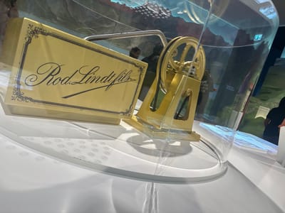 The image shows a gold-colored model of a Lindt & Sprüngli chocolate refiner, encased in a transparent cylindrical display case with a white top and base. The display case is located indoors, likely within the Lindt Home of Chocolate museum in Kilchberg (ZH), Switzerland, as evidenced by the Lindt branding on the model. The model features a wheel mechanism and a rectangular plaque bearing the words "Rod. Lindt." in ornate script. The environment around the display case includes illuminated surfaces and possibly projections or displays visible through the transparent case. Reflections and distortions are present on the display case surface, hinting at other displays, artifacts, or people nearby.
