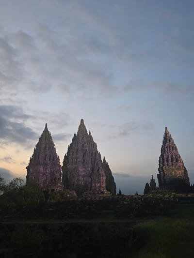 The image captures the Prambanan Temple complex in Bokoharjo, Indonesia, against a backdrop of a cloudy sky at dusk or dawn. Three main temples dominate the foreground, their stone structures and tiered roofs clearly visible. The lighting casts a soft glow on the temples, highlighting their intricate carvings. To the right, vegetation and some portions of other temple structures are visible.