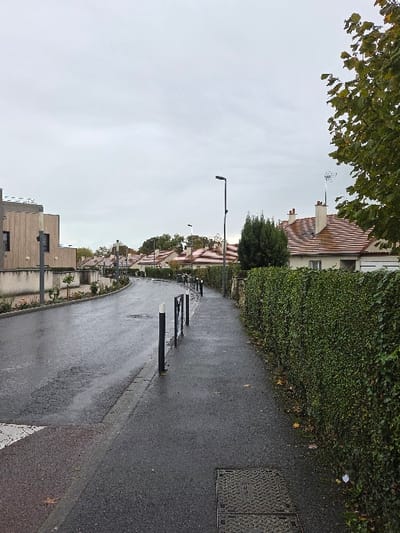 The image depicts a residential street scene in Blainville-sur-Orne, France, under an overcast sky. The asphalt road and sidewalk are visibly wet, indicating recent precipitation. The perspective is from the sidewalk, looking forward as the road curves gently to the left and uphill into the distance.
On the left side of the scene, a portion of a white-marked crosswalk is visible leading to the wet, dark asphalt road. Further along the left, a modern building with light-colored, possibly wooden, paneling and dark windows is partially visible. This building features a flat roof.
The right side of the image is dominated by a dense, tall green hedge lining the sidewalk. A rectangular metal utility cover is embedded in the wet sidewalk in the lower right foreground, with a small piece of white litter near the base of the hedge.
Separating the sidewalk from the road is a series of black bollards with white tops, spaced along the curb. Further down the street, a black metal barrier is also present. Several residential houses with light-colored facades and reddish-orange tiled roofs are visible beyond the hedge and bollards, extending into the midground. Some houses feature chimneys and television antennas.
Two black streetlights of a modern design are present, one prominently in the mid-right, and another further in the distance. A small, dense, dark green tree or bush is situated behind the prominent streetlight. Additional trees with green and some autumnal leaves are visible on the far right and in the background, interspersed among the houses and against the uniform light grey sky. No individuals are present in the scene.