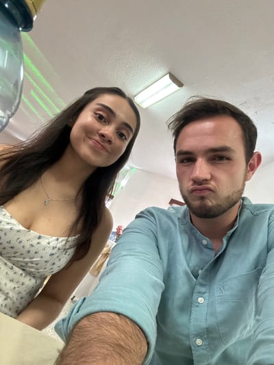 Two individuals, a man and a woman, are in close proximity, likely taking a selfie. The man is wearing a light blue button-down shirt. The woman is wearing a white floral print dress and a necklace. They appear to be indoors, possibly at a gathering or event, as suggested by the presence of other people and objects in the background. The location is Chipilo de Francisco Javier Mina (Chipilo), Mexico. Fluorescent lights are visible on the ceiling. A water dispenser is partially visible in the lower-left corner.