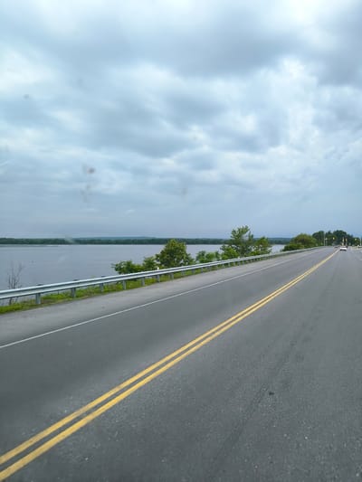 The image shows a road with double yellow lines, bordering a body of water with a tree-lined shore. The sky is overcast with heavy clouds. In the distance, there are a few cars on the road and a structure that appears to be a traffic signal. The scene is likely a highway or a rural road near a body of water. There are no people visible in the image. The image is taken from a moving vehicle, as indicated by the motion blur and perspective.