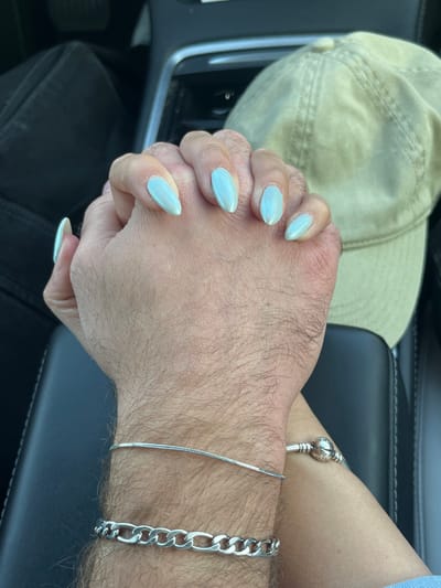 The image displays a close-up view of two individuals' interlocked hands and forearms within the interior of a vehicle. One hand, characterized by light blue, almond-shaped manicured fingernails, is positioned atop the other. The underlying hand and forearm are visibly hairy. The hairy wrist wears two silver bracelets: a thin snake chain and a thicker, flat link chain. The other wrist, less hairy, displays a silver Pandora-style charm bracelet with a spherical clasp. The background reveals black car upholstery on the left, a partial view of a dashboard or center console area, and a light-colored (beige/khaki) baseball cap resting on a surface in the upper right. A dark armrest or console is visible beneath the cap. The precise location within Rosh Haayin, Israel, cannot be determined from the visual information provided in the image.