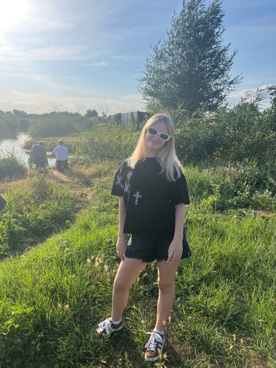 A young woman with blonde hair and white sunglasses stands in a grassy field, looking towards the camera. She is wearing a black t-shirt with a silver cross design and black shorts. She has on white and brown sneakers. In the background, a body of water is visible with two people fishing. The surrounding area is covered in tall green grass and reeds, with a large tree in the distance. The sky is blue with some clouds. The location is identified as Yanovo-Grushevskiy, Russia.