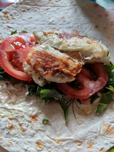 The image shows a close-up, overhead view of a chicken wrap being prepared on a flatbread. The wrap is filled with grilled chicken pieces, red tomato slices, and fresh green herbs, possibly dill and lettuce. A white sauce, likely mayonnaise or yogurt-based, is spread on the flatbread and also mixed with the filling. The flatbread itself appears to be lightly toasted, with some browned spots. The background is out of focus, suggesting the photo was taken indoors. The scene is presented in a food photography style, focusing on the appetizing ingredients of the wrap.
