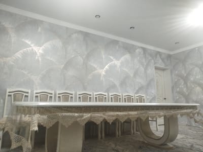 The image depicts a dining room in Petrovka, Kyrgyzstan. The room features a long, white dining table with a decorative lace tablecloth. A row of matching white chairs with upholstered seats is arranged around the table. The walls are covered in textured wallpaper with a light, shimmering pattern. The ceiling is white with recessed lighting fixtures. A doorway is visible on the right side of the room, leading to another area. The lighting in the room is bright, with a strong light source visible in the upper right corner, possibly from a window or a ceiling light. The overall impression is of a well-decorated and spacious dining area.