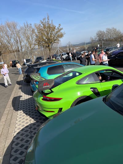 The image depicts an outdoor gathering in a parking area in Meuspath, Germany, under a clear blue sky. In the foreground, the dark green hood of a vehicle is partially visible. Immediately behind it is a bright lime green Porsche 911 GT3 RS, identifiable by its rear lights, side vents, and a large carbon fiber rear wing bearing a red "MANTHEY" logo. A white decal reading "@Kevinlabenski" is present on the Porsche's rear window, and an individual is seated in the driver's seat, holding a red beverage can.
To the left of the lime green Porsche, a metallic teal Volkswagen Golf R is parked, followed by a dark grey hatchback. A line of assorted parked vehicles, including several dark-colored cars and SUVs, extends into the midground. Approximately twenty individuals, predominantly men, are dispersed throughout the scene, either standing in groups, conversing, or observing the parked vehicles. On the far left, a young male in light-colored clothing walks away from the camera.
The ground surface in the foreground consists of interlocking concrete pavers, transitioning to asphalt further back. A chain-link fence runs across the mid-background, separating the parking area from a landscape of dormant trees and rolling hills. Strong shadows are cast, indicating direct sunlight from a high sun position.