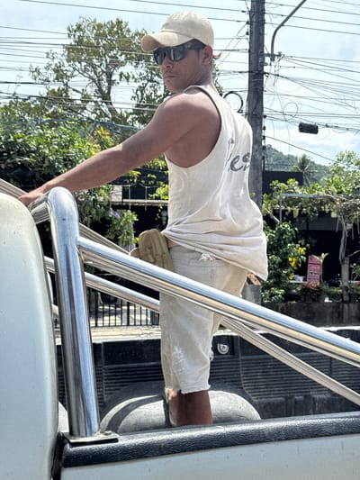 A man in a light-colored tank top, beige shorts, and a baseball cap stands in the bed of a pickup truck, looking over his right shoulder towards the camera. He wears dark sunglasses. A tan utility pouch is clipped to his belt. The truck bed contains a spare tire. Behind the truck, a utility pole with numerous wires is visible against a hazy sky. Lush green trees and foliage line a street, with hints of buildings and a distant hillside. The scene suggests a bright, sunny day in a tropical or subtropical urban environment, likely Baan Tha Po, Thailand, given the context.