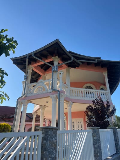 The image captures a large, multi-story house with distinct architectural features, situated in Petrovac-na-Mlavi Municipality, Serbia. The house is painted a vibrant pink and features a prominent balcony supported by numerous white Corinthian columns. The balcony is adorned with an ornate white railing. The roof is dark and appears to be tiled, with visible gutters. Two arched windows are visible on the upper level. In the foreground, a white picket fence with stone-capped pillars separates the property from the street. A dark-leaved shrub is planted in front of the house, partially obscuring the ground floor. To the left of the house, another building with a red-tiled roof is partially visible, as well as a small green shrub. The sky is a clear, bright blue. The scene is bathed in sunlight, suggesting a clear day. No people or animals are visible in the image.