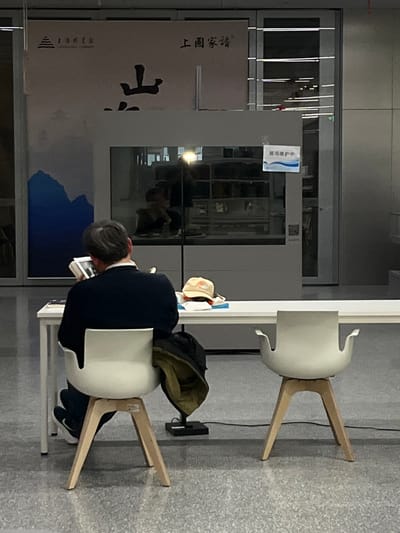 The scene depicts an interior view of the Shanghai Library. A male individual, with graying hair, is seated with his back to the camera at a white table, reading an open book. He is wearing a dark long-sleeved garment and dark pants; a dark green jacket is draped over his white chair, which features wooden legs. To his right, an identical empty white chair with wooden legs is positioned at the same table. On the table, near the reader, rests a light-colored baseball cap with a red insignia and a small stack of white papers or a booklet.
In the background, a large beige display panel features the "SHANGHAI LIBRARY" logo and Chinese characters, including "上海图书馆" (Shanghai Library), with a blue and white abstract mountain design below. Adjacent to this, a glass partition reflects the partial image of a person and a bright light source. A small white sign with Chinese text, likely indicating "Exhibition under maintenance," is affixed to the glass, along with a QR code. The floor is light gray and tiled.