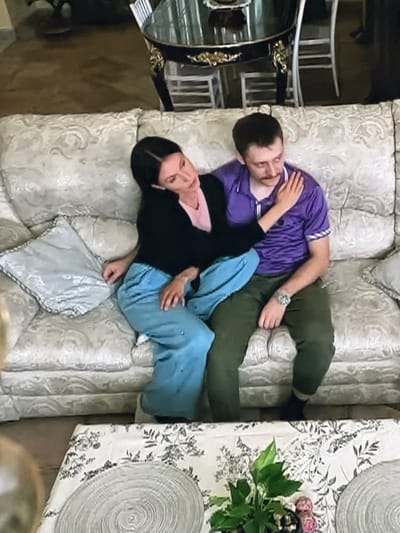 A couple is seated on a light-colored, ornate couch. The woman, with dark hair, is leaning into the man and has her arm around his shoulder. She is wearing a black cardigan over a pink top and light blue pants. The man, with short brown hair and a mustache, is wearing a purple polo shirt and green pants. He has his arm resting on the back of the couch, and his other hand is partially visible in the frame. On the couch are decorative pillows, one of which is light gray. In the foreground, a table is visible with a floral tablecloth and placemats, and a small potted plant. The background shows parts of a dining area with a dark, elaborate table and chairs. The scene appears to be indoors, likely a home, in Dondușeni, Moldova.