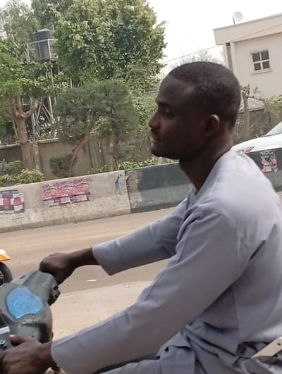 A dark-skinned male, with short dark hair, is positioned in profile, facing right, in Darmanawa, Nigeria. He wears a light grey long-sleeved garment. Both hands grip the grey handlebars of a motorcycle or scooter, the control panel of which features a light blue, possibly worn or applied, surface. In the midground, a concrete roadside barrier, partially obscured by faded posters or graffiti, separates the subject from a dirt road. The background reveals a light-colored building with a square window and a rooftop satellite dish. Dense green foliage, including various trees and a black water tank on a metal stand, is present. A white vehicle is partially visible on the right side of the road behind the barrier. The scene is illuminated by daylight.