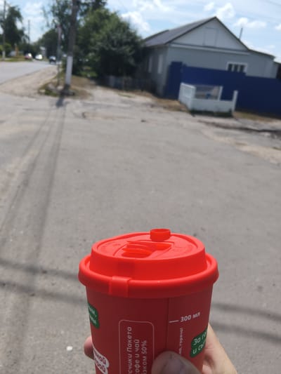 The image shows a red coffee cup with a red lid held by a person's hand. Text on the cup is visible, including "300 мл" and some Russian phrases. The background suggests an outdoor urban environment. In the distance, there is a road with tire tracks, a sidewalk, a utility pole, trees, and a building with a blue fence. The scene appears to be a street in Mokshan, Russia, based on the presence of Russian text on the cup and the characteristic architecture of the building. The sky is partly cloudy.