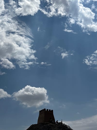 The image shows a view of the Jiayu Pass in Jiayuguan, China, under a bright blue sky with scattered white clouds. The focus is on the fortress structure atop a rocky outcrop in the foreground, silhouetted against the sky. The fortress is a small, square tower with crenellated battlements, made of reddish-brown material. Several small human figures are visible standing on a walkway or platform to the right of the fortress, also in silhouette. The overall impression is of a clear, sunny day with the historic site prominently displayed.