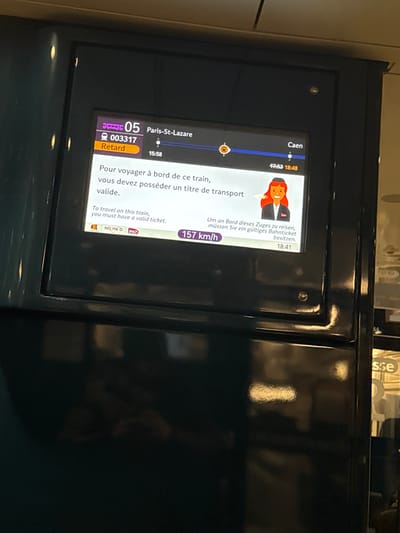 The image shows a train information display. The train is number 05, and it's delayed ("Retard"). It's traveling from Paris-St-Lazare, scheduled to depart at 15:58, and heading towards Caen, with an arrival time of 18:48. The train's current speed is 157 km/h.  The display advises passengers in French, English, and German that they must have a valid ticket to travel on the train. A cartoon of a train conductor is displayed.