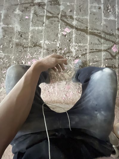 The image shows a person's legs and arms, sitting on a rough, debris-strewn floor. The floor appears to be concrete with visible grid lines marked in white. Scattered across the floor are small pieces of pink material, possibly plaster or paint chips. The person is wearing dark pants that are faded and stained, and a light-colored shoe is visible on the right foot. A white wire, possibly from headphones or a device, hangs down between the legs. The background suggests an unfinished or construction site environment. There is no visible indication of the specific location within Jihlava, Czechia.