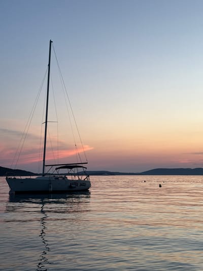 The image depicts a serene sunset scene on the water, likely in Grad Šibenik, Croatia. A sailboat, named "RAZZLE DAZZLE" as indicated by visible text on its hull, is moored in the calm water. The boat is facing towards the right of the frame, its mast and rigging silhouetted against the colorful sky.
The sky transitions from a pale blue at the top to soft shades of pink and orange near the horizon, suggesting the time of day is sunset. The water reflects these colors and the boat, creating a shimmering effect. A distant shoreline with a dark silhouette of hills or land is visible on the right side of the image, separating the sky and water. The overall mood is peaceful and tranquil, enhanced by the gentle ripples on the water's surface. There are no people visible in the frame. The weather appears calm, with no indication of wind or rough seas.