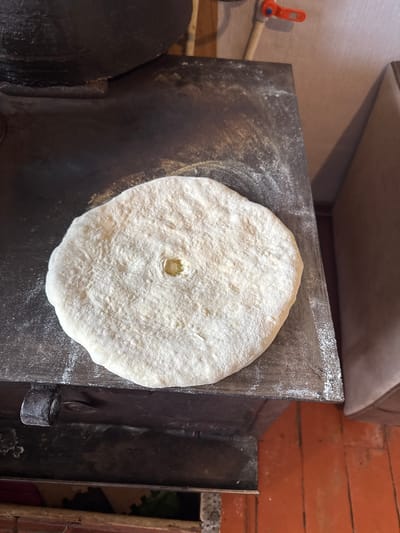 A flatbread, dusted with flour, rests on a dark, worn metal surface. The surface appears to be the top of a wood-burning stove, as evidenced by the black, sooty material and a metal handle on the left side. The flatbread has a small, indented circle in its center. The background shows a textured, light-colored wall and a portion of a brown upholstered object, possibly a chair or sofa. The floor below is made of reddish-orange tiles. The scene is likely in a kitchen or cooking area. The image does not provide enough information to determine the exact location in Tandzia, Georgia.