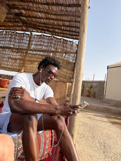 An adult Black male with dark skin, wearing sunglasses and a white t-shirt, sits outdoors on a patterned rug. He has earbuds in his ears and is holding a silver smartphone in his right hand. He is wearing a green-stone ring on his left index finger and a silver ring on his right index finger. His left arm is crossed over his chest. He is seated in a shaded area with a thatched roof supported by a wooden post. The ground is sandy. In the background, there is a tan building, a woven fence, and potted plants. The location is Agafay, Morocco.