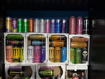 The image shows shelves stocked with a variety of canned energy drinks. The drinks are arranged in rows, with different brands and flavors visible, including Red Bull and Monster Energy. Some of the cans are brightly colored, featuring prominent logos and branding. The shelves appear to be made of cardboard or a similar material, suggesting a retail or storage environment. The background is dark, possibly a wall or the interior of a cabinet. The image does not provide any information about Zhabokrat, Bulgaria, or any people, actions, or interactions within the scene.