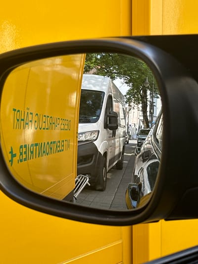 In a close-up shot of a car's side mirror, the reflection reveals a traffic scene on a city street in Gütersloh, Germany. The mirror is mounted on a bright yellow vehicle, which occupies a significant portion of the foreground and left side of the image. Reflected in the mirror, a white delivery van is visible, partially obscuring a dark-colored car behind it. The van has its headlights on, and the road ahead is lined with trees and buildings. Green text is printed in reverse on the yellow vehicle, which reads "DIESES FAHRZEUG IST EIN ELEKTROFAHRZEUG" and "MIT ELEKTROANTRIEB".