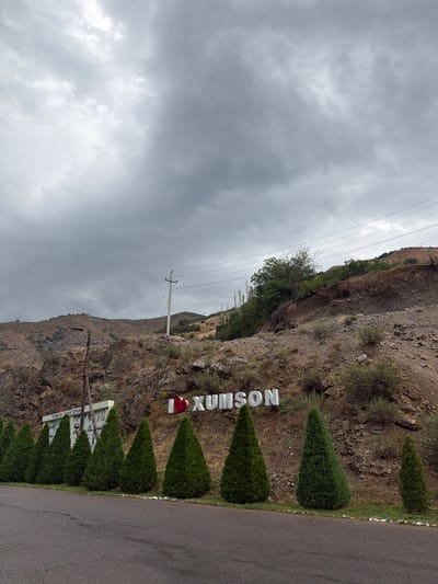 The image captures a landscape view of Khumsan, Uzbekistan. In the foreground, a paved road runs parallel to a row of neatly trimmed evergreen trees. Behind the trees, a rocky, arid hillside rises. On the hillside, large white letters spell out "I ❤️ XUMSON," with a red heart replacing the word "love." To the left of the letters, a white building with colorful murals is visible, with Cyrillic text above it that reads "Хумсон санатория" (Khumsan Sanatorium). A power line with poles and wires runs across the middle ground, crisscrossing the scene and extending towards the background hills. The sky above is overcast with heavy, gray clouds, suggesting a potentially stormy or gloomy day. The overall impression is of a scenic, possibly rural location with a clear identifier of the town's name.