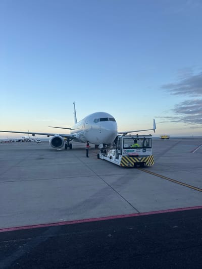 The image shows the front of a Boeing 737 aircraft connected to a pushback tug, likely at Tenerife South Airport in Granadilla de Abona, Spain. The tug is yellow and black with “Menzies” branding. A person in a high-visibility vest is inside the tug. The aircraft is white and silver with visible engine nacelles. Yellow and black guiding lines are painted on the tarmac. In the background, other aircraft and airport vehicles can be seen along the runway. The sky is a clear blue with some clouds.