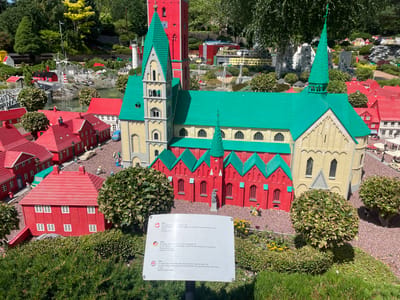 The image depicts a miniature model of Ribe, Denmark, located within the Legoland Billund theme park. The central subject is a detailed Lego replica of Ribe Cathedral, a prominent red and yellow brick structure with a striking green roof and steeple. Surrounding the cathedral are smaller Lego buildings, also constructed with vibrant red roofs and walls, creating a charming, scaled-down townscape. A sign in the foreground provides information about Ribe in multiple languages, mentioning its founding in approximately 700 AD and its historical flood events. Several Lego figures are scattered throughout the scene, engaged in various everyday activities, adding a sense of life to the miniature world. Lush green foliage and trees frame the Lego structures, suggesting an outdoor setting bathed in sunlight. The overall impression is one of a meticulously crafted and immersive Lego village, showcasing historical architecture in a playful and engaging manner.