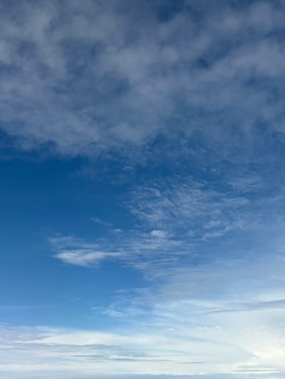 The image depicts a daytime sky over Amora, Portugal, entirely filled with atmospheric elements. The upper third of the frame shows a medium-blue sky with scattered, fragmented white and light-grey cloud formations, appearing as altocumulus or cirrocumulus. The middle section transitions to a brighter blue, featuring sparser, wispy white clouds indicative of cirrus formations. The lower third displays a paler blue sky leading to a more continuous, denser band of bright white cloud cover near the bottom of the frame, suggesting a lower cloud layer or haze. No ground features, objects, people, or actions are visible within the scene.