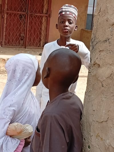 This bright daylight outdoor photograph captures three children interacting in what appears to be a casual setting in Fanisau, Nigeria.
The central figure is a young boy, approximately 8-12 years old, facing the camera with a distinct expression. He is wearing a white long-sleeved tunic and a patterned Kufi cap with white, red, and black geometric designs. His lips are puckered in a pout, and he points with his right index finger, seemingly engaging with one of the other children.
To his left, partially visible from the back, is a girl wearing a white hijab or headscarf, which has small, subtle embellishments. Her face is turned away from the camera, and she is holding a light-colored, possibly plastic, bag or object.
On the right side of the frame, another boy is visible from the back. He has a shaven head and is dressed in a dark brown or grey long-sleeved tunic. He stands close to a textured, possibly stucco or mud, wall or pillar that occupies the right edge of the image.
In the background, behind the central boy, there is a building featuring a reddish-brown metal gate or door with a grid-like pattern. The ground appears to be unpaved dirt or sand. The lighting suggests it is daytime. There is no visible text in the image.