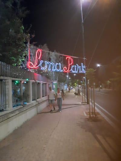 In Pesaro, Italy, a neon sign reading "Luma Park" illuminates the night. The sign is composed of red neon for "Luma" and blue and green neon for "Park," strung across wires suspended by poles. Beneath the sign, a sidewalk is bordered by a low wall and a fence adorned with small, multicolored lights. A paved pathway stretches into the distance, with trees planted along the edge of a street to the right. Two individuals, a man and a woman, walk hand-in-hand on the sidewalk in the foreground. The man wears a white t-shirt and shorts, while the woman is in a white top and dark pants. Further down the sidewalk, other pedestrians and a bicycle are visible. The scene is set at night, with streetlights and the neon sign providing illumination.
