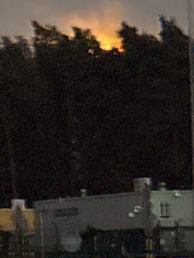 The image captures a dimly lit exterior scene, possibly at dusk or dawn, with a prominent Amazon logo visible on a building in the foreground. The sky behind a dense line of dark trees is illuminated by a bright, diffused orange light, suggestive of the sun. The building appears to be a commercial or industrial structure, with multiple sections and a flat roof. Some details of the building's facade, such as windows and architectural elements, are discernible despite the low light conditions. The overall impression is one of a quiet, perhaps isolated, location with a focus on the presence of the Amazon facility.