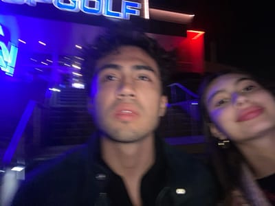 A slightly blurred photo shows two individuals at night in front of a "Topgolf" establishment. The man is centered, looking upwards with a dark jacket and short dark hair. A woman appears on the right side of the frame, smiling towards the camera. Stairs and various blue and red lights can be seen behind them.