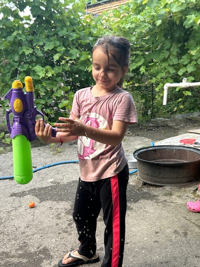 A young girl is standing outdoors, holding a purple and green water gun. She is wearing a pink t-shirt with a unicorn graphic and black pants with a red stripe. Her hands are outstretched, and small water droplets are visible in the air around her and on her clothing, suggesting she may be in the process of playing with the water gun or has just used it. The background features lush green foliage, likely a vine-covered wall or trellis, and a concrete ground surface speckled with water. A large, dark-colored metal basin is positioned to the right of the girl, and a blue hose lies on the ground. To the far right, a pink clog is visible. The scene appears to be in a residential yard or outdoor play area. The lighting suggests it is daytime, possibly overcast. The environment appears to be urban, consistent with a location in the city of Prokhladny, Russia.