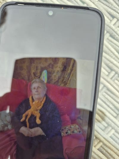 A framed photograph, possibly a print of an old photograph, is displayed on a phone screen. The photograph depicts an elderly woman with gray hair, wearing a dark garment and an orange scarf. She is seated on a red sofa, holding a small orange stuffed animal. A patterned curtain is visible in the background. The phone itself is partially visible, resting on a woven surface. There are some specks of dust or debris on the phone screen and the photograph. The location of Carrick-On-Shannon, Ireland, is not indicated by any visual elements in the image.
