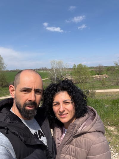 A medium shot shows a man and a woman outdoors on a sunny day. The man is positioned in the foreground, with the woman slightly behind and to his right. The man has a shaved head, a dark beard, and is wearing a gray shirt and a black jacket. The woman has dark, curly hair and is wearing a light brown quilted jacket. Both are looking toward the camera with slight smiles.

The background includes a blue sky with scattered clouds, green grass, trees, and a dirt road. The setting appears to be a rural or semi-rural area. The location in Slanotran, Bulgaria, is not identifiable from the image.