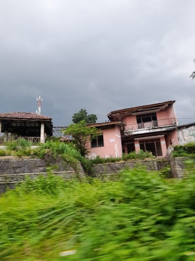The image depicts an overgrown, possibly abandoned, area in Kecamatan Prigen, Indonesia, under an overcast sky. Two dilapidated buildings or structures, likely old houses or a resort, are the central subjects.
The main building on the upper right side (when viewed correctly) is a two-story structure with faded reddish-pink walls and a traditional tiled roof that shows signs of decay, including missing tiles and damaged eaves. Its windows are dark and appear broken or empty. Below and to the left of this building is another smaller, single-story structure with a similar roof style, also appearing abandoned.
Lush, vibrant green grass and other vegetation heavily overgrow the area, particularly on the right side of the image, suggesting neglect and the passage of time. This foreground vegetation appears somewhat blurry, possibly indicating motion blur from the camera or a shallow depth of field. Stone retaining walls or terraces are visible between the buildings and the overgrown slope. In the distance, partially obscured by the structures, a communication tower (like a cell tower) stands against the gray sky, providing a modern contrast to the decaying architecture.
The sky is uniformly gray and cloudy, suggesting an overcast day, possibly signaling impending rain or simply a lack of direct sunlight. There are no people visible in the scene. The overall impression is one of abandonment and nature reclaiming man-made structures. No visible text is present.