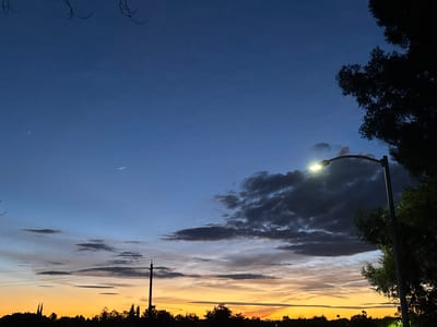 This media file captures a twilight scene in Sacramento, United States. The image features a dusky sky transitioning from a deep blue at the top to vibrant oranges and yellows near the horizon. Wispy clouds are scattered across the sky, some dark and brooding, others lighter and streaky, adding texture and depth.

A street lamp with a bright, illuminated bulb stands prominently on the right side of the frame, its light cutting through the fading daylight. Silhouetted against the colorful sky are the dark shapes of trees, power lines, and a distant treeline, suggesting an urban or suburban environment. The branches of a tree are also visible in the upper left corner, adding a natural element to the man-made structures.

The time of day is clearly evening, as indicated by the sunset colors in the sky and the activated street lamp. The weather appears clear but with scattered cloud cover, contributing to the dramatic sky. There are no people or overt activities depicted, creating a sense of calm and quiet observation. The overall mood evoked is serene and picturesque, highlighting the beauty of the natural light at dusk. No visible text is present in the image.