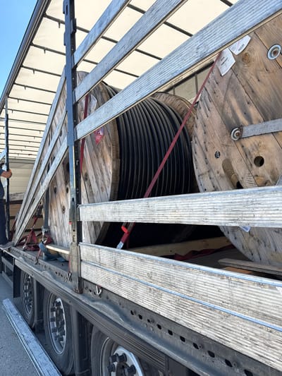 The image captures a close-up, eye-level view of a flatbed truck loaded with large wooden spools of black cable. The spools are positioned lengthwise on the truck bed and secured with red and blue straps. The truck's tires and undercarriage are visible in the lower portion of the image, suggesting an outdoor setting on pavement. The spools themselves appear weathered, made of rough wooden planks held together with metal fixtures. Some of the spools have paper labels attached. The truck is covered with a white tarp, and metal support beams are visible along the sides. A person wearing dark clothing is partially visible on the left side of the truck, appearing to be standing near the load. The sky above is a clear blue, indicating daylight. The scene is devoid of any distinctive landmarks or elements that would specifically place it within the city of Smyadovo, Bulgaria.
