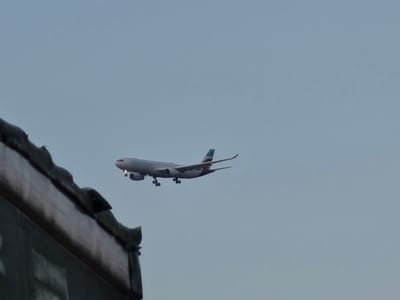 An airplane, identified as a Lufthansa Airbus A330-300, is captured in mid-flight against a clear sky. The aircraft is descending, indicated by its landing gear extended. The front of the airplane has its navigation lights illuminated. In the foreground, a dark, textured building structure occupies the left portion of the frame, appearing to be a rooftop or part of a larger building. The airplane is positioned to the right of the building structure. The overall scene suggests an approach to Frankfurt Airport (FRA), which is located in Kelsterbach, Germany.