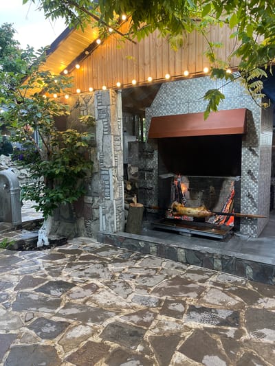 The image shows an outdoor cooking area in what appears to be a patio or garden setting. A whole animal, likely a pig, is being roasted on a spit over an open fire. The cooking area is constructed of stone and brick. There is a canopy above with string lights hanging from it. The ground is paved with stone tiles. Plants and trees are visible around the cooking area. The image does not contain evidence to confirm that it is taken in Miatli, Russia.