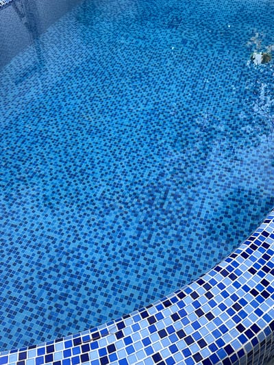 The image depicts the interior of a swimming pool in Khujakent, Uzbekistan. The pool is filled with clear blue water, revealing a tiled mosaic bottom composed of small, square blue tiles in varying shades. The edge of the pool is visible in the foreground, also lined with a mosaic of blue and white tiles. The lighting suggests an outdoor setting, with some reflections on the water's surface hinting at sunlight and possibly overhead foliage or structures. There are no people or animals visible in the image, and no actions or interactions are taking place. The environment is solely focused on the architectural elements of the swimming pool.