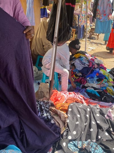The scene depicts an outdoor market in Sabon Gari South, Nigeria. In the immediate foreground on the left, a person, identifiable by a light purple long-sleeved shirt and a ring on the right ring finger, holds a large roll of dark purple fabric with a subtle repeating pattern. In the mid-ground, positioned slightly right of center, another individual is seated on a light blue plastic stool. This person wears a black beanie with white markings, a white long-sleeved shirt, and white pants, and is actively sifting through a substantial, multi-colored pile of textile goods, including various fabrics and garments, laid directly on the light brown dirt ground. This pile contains items with polka dots, floral patterns, and solid colors such as red, blue, orange, and green. A vertical, partially rusted metal pole stands prominently in the mid-ground, from which a large black plastic bag hangs. To the left of this pole, a rough, light brown stone rests on the ground. In the background, several other individuals are visible, including one seated on another light blue stool wearing a beige garment, and others standing or moving amongst stalls displaying various hanging clothing items like dresses and tops in blue, pink, and black. The overall environment is open-air, with scattered debris on the ground and various market structures suggested in the distance.