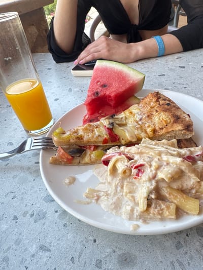 A close-up, eye-level shot shows a plate of food, a glass of orange juice, and a person's arm on a speckled grey outdoor table. The plate contains two slices of pizza and a portion of pasta with a creamy sauce, red pepper, and what appears to be mushrooms and cheese. The pizza has a slightly burnt crust. Next to the plate is a tall glass filled with orange juice. A fork rests on the left edge of the plate. A woman's arm, adorned with a blue wristband, is visible in the upper right quadrant. The background suggests an outdoor setting with a wooden table and some foliage. The overall scene appears to be a meal at a restaurant or cafe. The image's context suggests it was taken in Agios Nikolaos Municipal Unit, Greece, likely at a dining establishment near the coast given the outdoor setting.