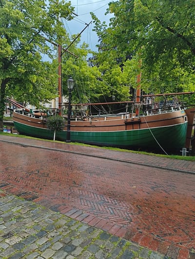 The image shows a historic sailing ship docked in a canal in Papenburg, Germany. The ship has a brown hull with a green waterline. It is surrounded by lush green trees, and a cobblestone path runs along the canal. A black lamppost stands near the ship. The sky is overcast, suggesting a cloudy day. The overall scene evokes a sense of old-world charm and maritime heritage.