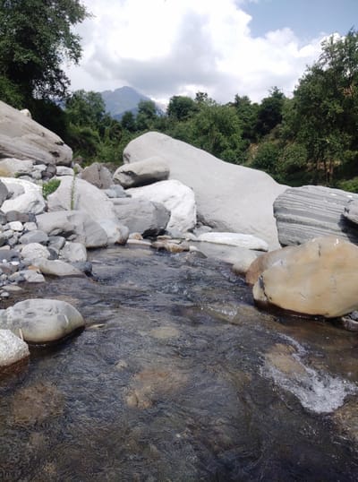 The image depicts a natural landscape, likely in the city of Baragran, India. In the foreground, a shallow, clear stream flows over a bed of rocks. The water is turbulent in some areas, creating small splashes and ripples. The stream banks are lined with a variety of stones and boulders, ranging in size from small pebbles to large, smooth rocks.
Behind the stream, the terrain rises, with lush green trees and dense foliage covering the slopes. In the background, a majestic mountain peak is visible, partially shrouded by clouds. The sky above is a mix of blue and white, with fluffy clouds scattered across it. The overall scene evokes a sense of tranquility and the raw beauty of nature. No people are visible in the image.