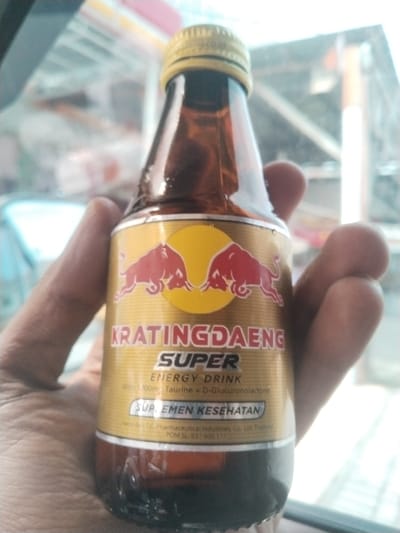 A person's hand holds a small brown glass bottle of Kratingdaeng Super Energy Drink. The bottle has a gold label with a red bull graphic. The background is blurred and shows what appears to be an indoor area with bright lighting, possibly a store or a vehicle interior. The location in the city of Reuhattuha, Indonesia is not specified in the image.