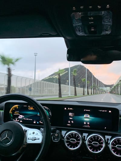 The image depicts a view from inside a Mercedes-Benz vehicle traveling on a multi-lane road in Ksar El Majaz, Morocco. The perspective is from the driver's or front passenger's seat, looking forward through the windshield.
In the foreground, the vehicle's interior is visible. The multi-function steering wheel, bearing the Mercedes-Benz logo, is prominent on the lower left. To its right, the digital instrument cluster shows a speed of "110 km/h", an outside temperature of "21.5°C", and a remaining distance of "24.6 km". A central infotainment screen displays various options, including "Maison" (Home), "Recherche" (Search), "Store", and the time "17:27". Below this screen are circular air vents with illuminated rings. An overhead console, located above the rearview mirror, contains "SOS" and "i" call buttons along with interior lighting controls. The rearview mirror reflects a blurred portion of the vehicle's ceiling and a dark shape that may be a headrest or occupant's head.
Outside the vehicle, a paved road, appearing damp, stretches into the distance. Numerous palm trees line both sides of the road. A tall, light-colored barrier or sound wall runs parallel to the left side of the road. Streetlights are regularly spaced along the roadside. In the background, a prominent, dark, rocky mountain is visible to the right. The sky is overcast, with a soft orange-pink hue near the horizon on the right, indicative of late afternoon lighting. The vehicle is in motion.