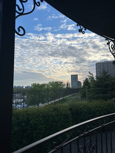 The image shows a view of buildings and a sky with scattered clouds framed by a wrought iron structure. Tall modern buildings are visible in the background, partially obscured by trees and a black fence. A low building with multiple square windows is also visible. The sky transitions from blue to a more muted, hazy light towards the horizon. The wrought iron frame is dark and ornate, with decorative swirls. The scene appears to be captured from an elevated position, possibly from a balcony or window. The location is Kuleshovka, Russia.
